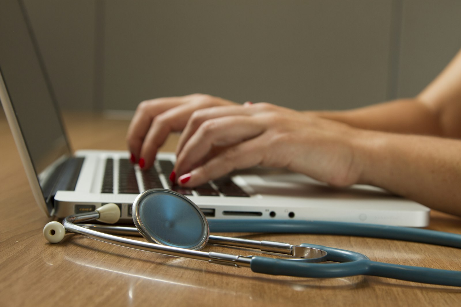 Hands typing on a laptop beside a stethoscope on a clinician's desk